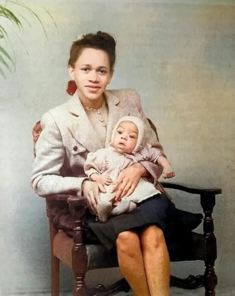 Lucille Jeter holding her baby son, Jimi Hendrix, while seated on a wooden chair against a nostalgic backdrop.