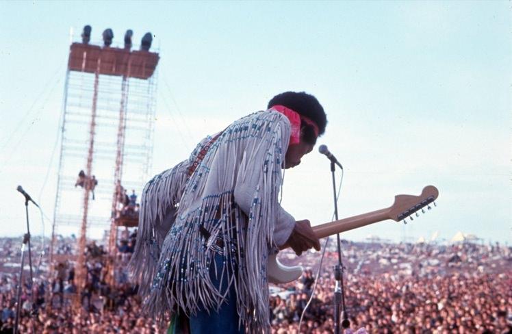 Jimi Hendrix is on stage at Woodstock, playing his guitar with a sea of spectators in the background.