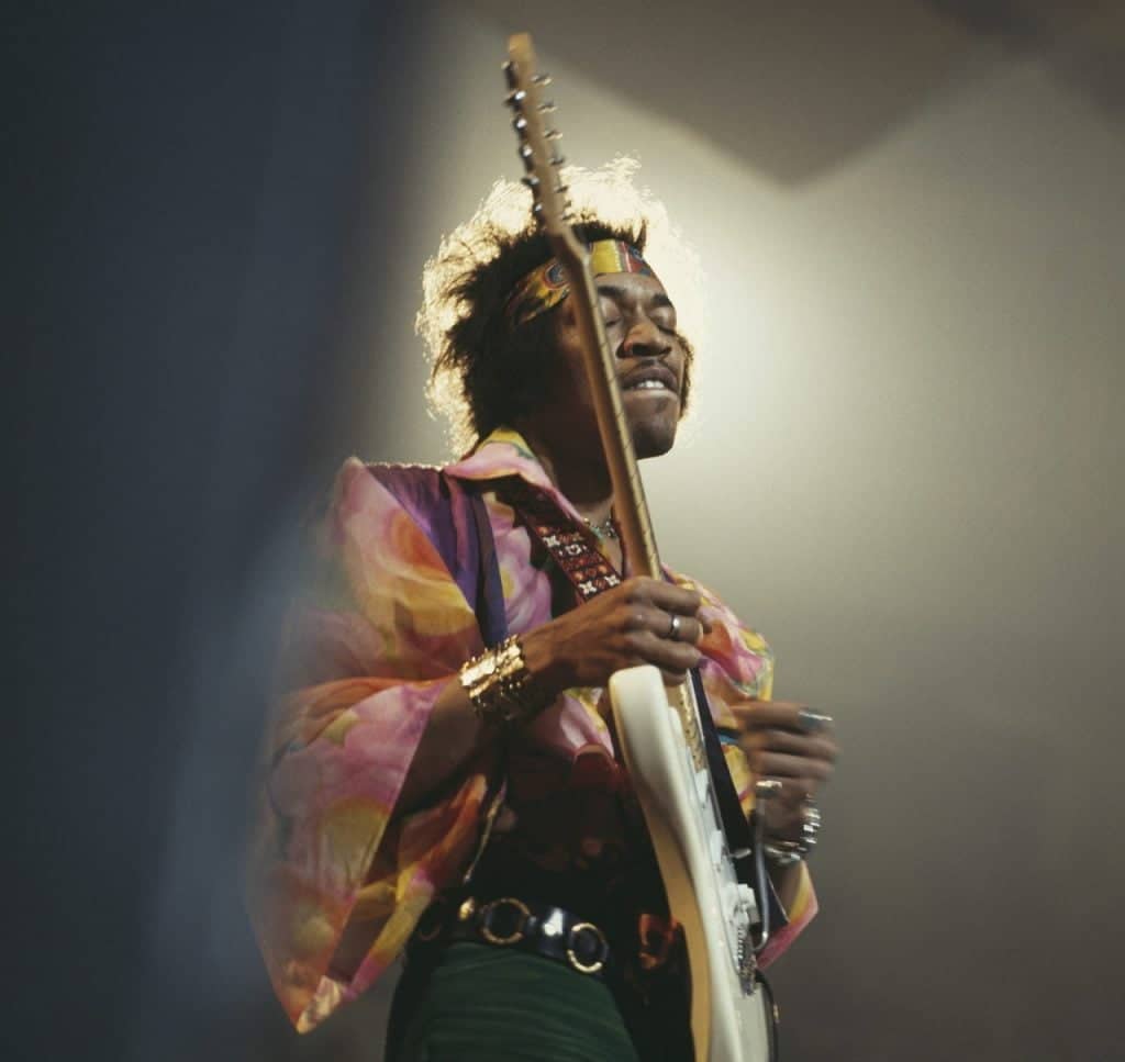 Jimi Hendrix playing his guitar on stage at the Royal Albert Hall, wearing a vibrant multi-colored shirt and a headband.