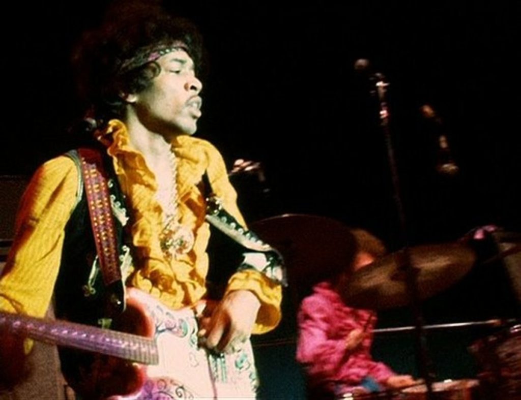 Jimi Hendrix on stage at the Monterey Pop Festival, wearing a vibrant yellow shirt and playing his fender guitar.