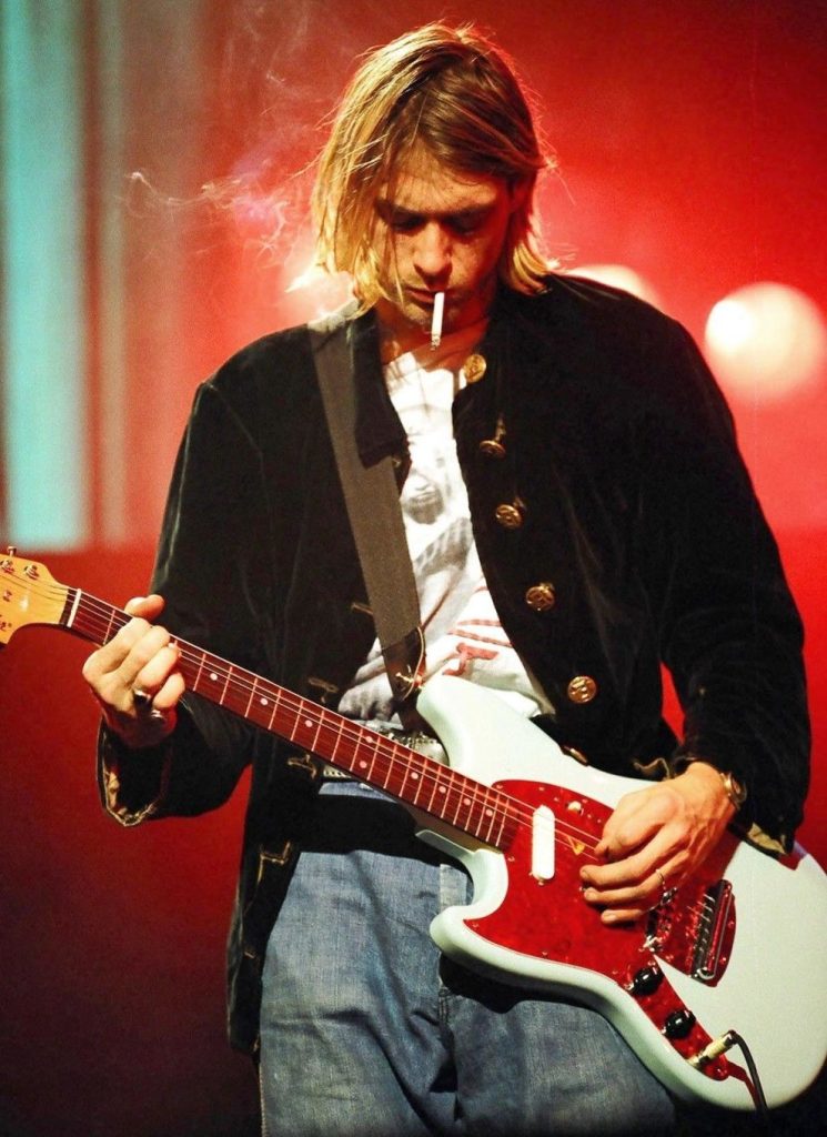 Kurt Cobain intensely playing a red guitar with a cigarette in his mouth, highlighted by stage lighting.