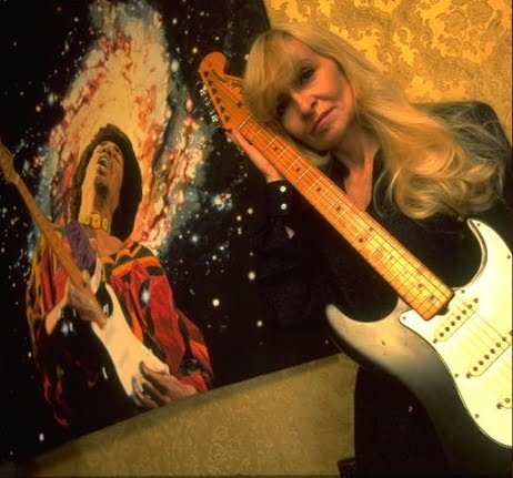 Monika Dannemann posing with Jimi's black Stratocaster beside a captivating painting of Jimi Hendrix.