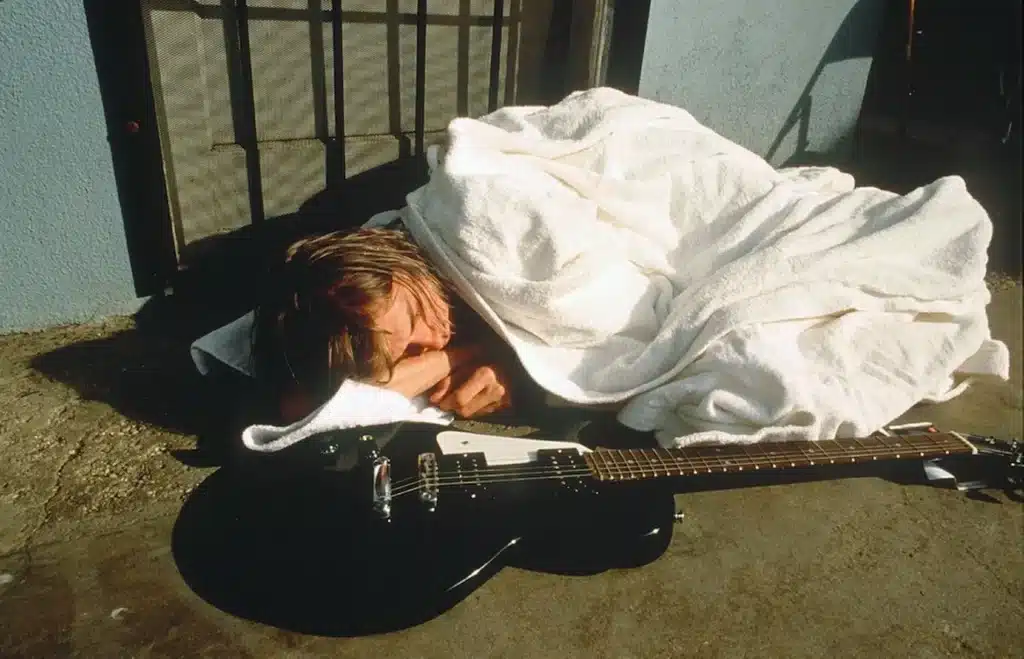 Kurt Cobain lying down on the floor with a blanket, accompanied by his guitar.